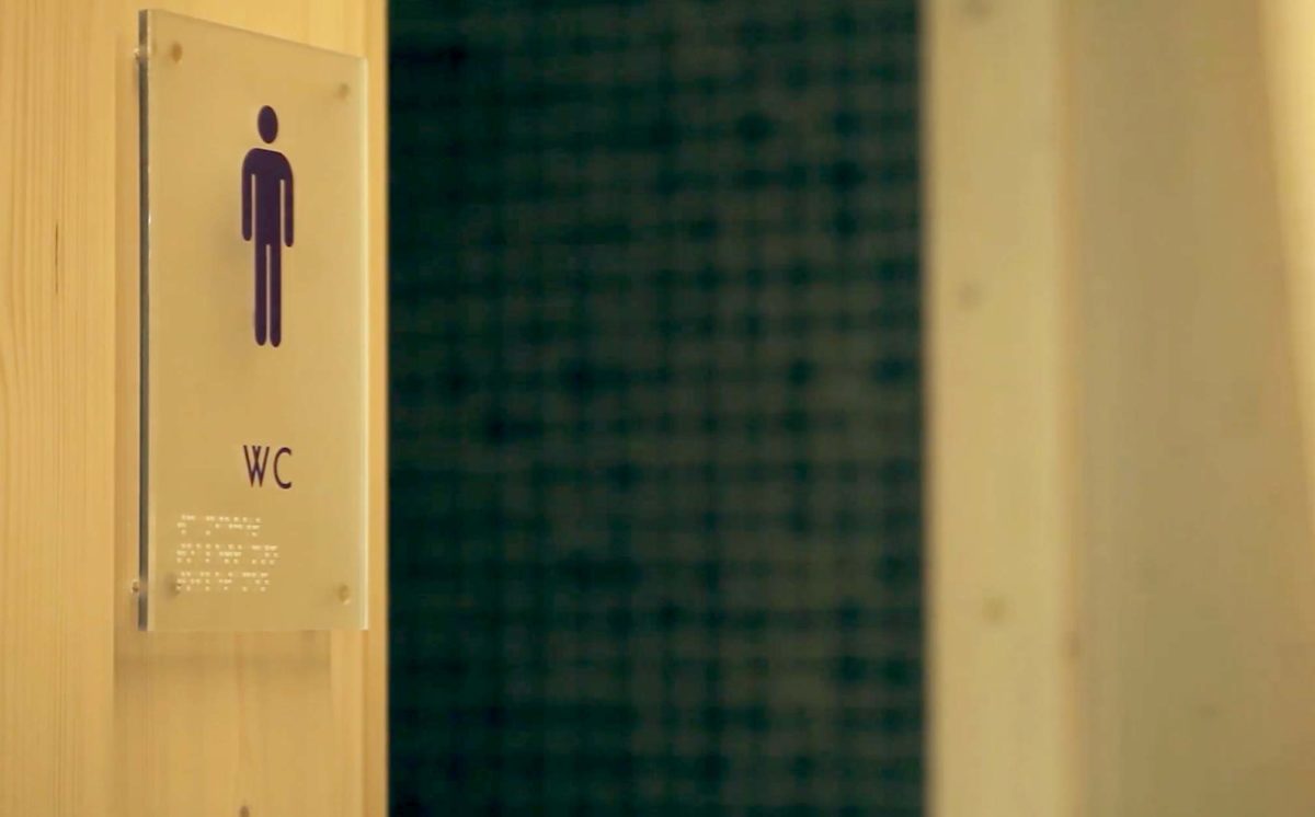 Miesten WC:n sisäänkäynti. Läpinäkyvässä kyltissä on symbolinen mieshahmo. Kyltissä lukee WC ja kyltti on merkitty myös pistekirjoituksella.