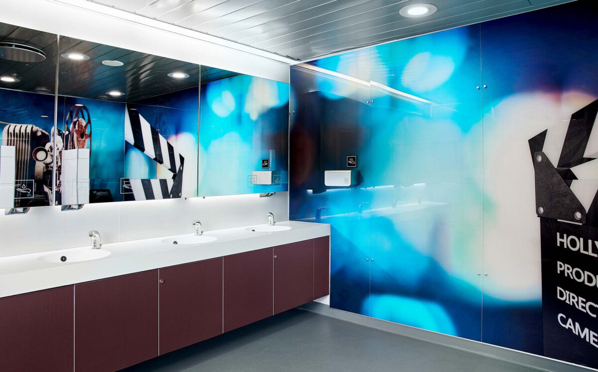 Novosan referenssi. Kauppakeskus Sellon miesten-WC. Vessassa on neljä valkoista käsienpesuallasta ja rusehtavan punaiset allaskaapit. Sinisen sävyisessä seinäpaneelissa näkyy elokuva-aiheinen kuva.