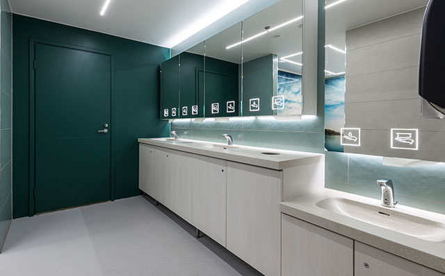 Vihreän sävyisessä WC-tilassa on kaksi käsienpesupaikkaa normaalikorkuisella tasolla ja yksi lavuaari alemmalla tasolla. Alakaappien ovet ovat vaaleat ja allastasot vaalean harmaat. Hanojen yläpuolella on Novosan peilikaapit, joissa on opasteet käsipaperille ja saippualle.
