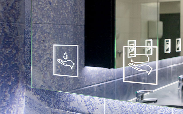 Novosan Pala-peilikaappi asiakas-WC:ssä. Peilikaapin peilissä on ohjaavat symbolit: käsisaippua- ja paperi. Peilistä heijastuu kuva toisesta peilikaapista ja hanoista. Vessan seinäpinnan kuviointi on rosoinen ja sinisen sävyinen.