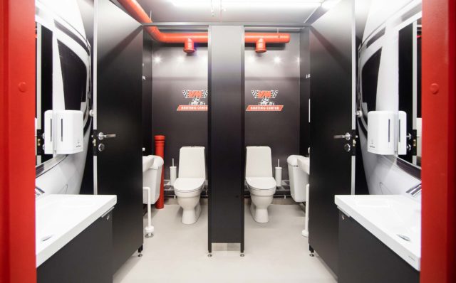 Mustavalkoisessa WC-tilassa on taustalla kaksi eriötä ja WC-pönttöä. Eröiden ovet ovat auki. Vastakkaisilla seinillä näkyy kaksi allaskalustetta ja niiden yläpuolella on kaarevat peilit. Altaiden viereen, seiniin on kiinnitetty saippua-annostelijat ja vaatekoukut. Punaiset ovenkarmit reunustavat kuvaa.