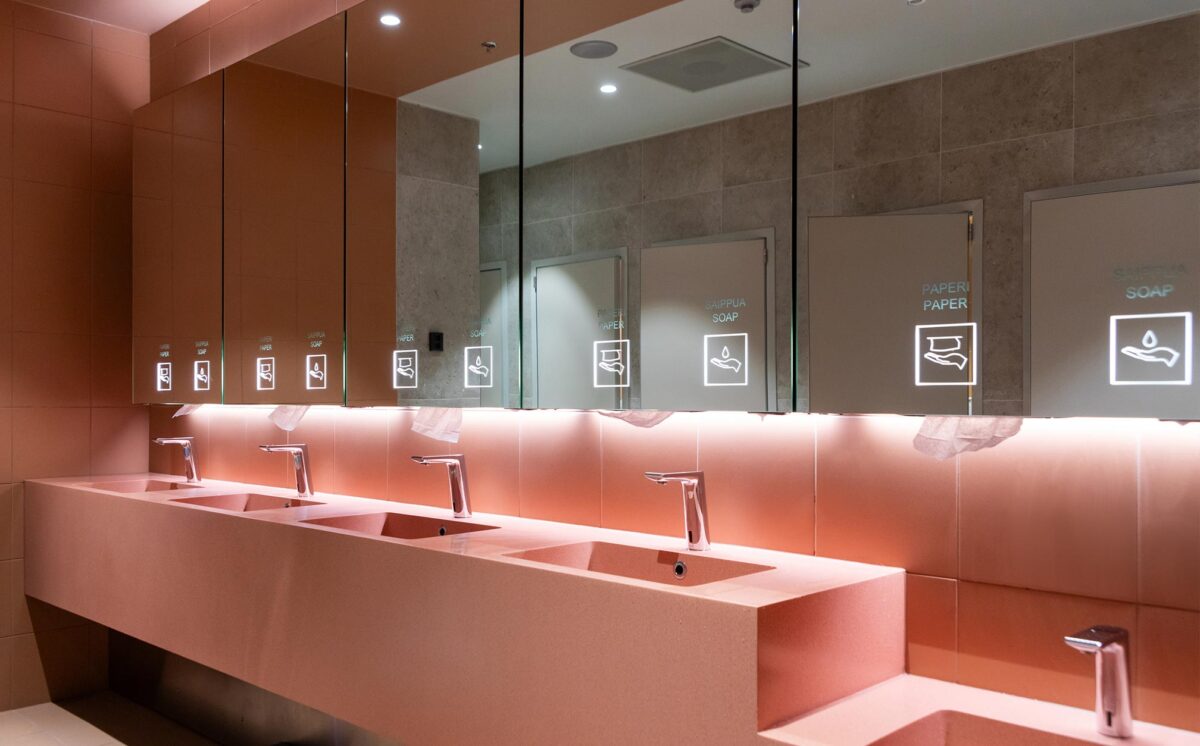 Vaaleanpunaisen sävyinen WC-tila, jossa on julkiseen tilaan sopiva peilikaappi Pala, allaskaluste, lavuaarit ja hanat. Pala-peilikaappi on valaistu ylä- kuin alalaidoista ja peilipinnassa näkyvät myös valaistut opasteikonit: paperi ja saippua.