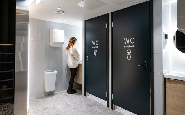 Novosan, WC-tilasuunnittelu. Näkymä siististä ja kauniisti valaistusta Sampokeskuksen WC-tilasta. Nainen pesee käsiään vasemman puoleisella pesualtaalla. Vessassa on keskellä kaksi WC-koppia yksi naisille, toinen miehille.
