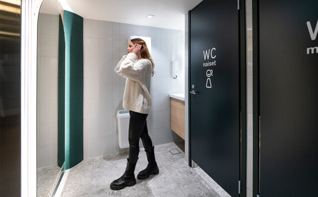Novosan, WC-tilasuunnittelu. Näkymä siististä ja kauniisti valaistusta Sampokeskuksen WC-tilasta. Nainen laittaa hiuksiaan kokovartalopeilin edessä. Vessassa on kaksi WC-koppia yksi naisille, toinen miehille.