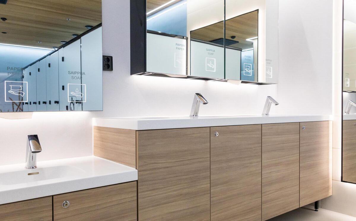 Tyylikäs julkinen saniteettitila, jossa on automaattihanat, puupintaiset allaskaapit ja valaistut peilit. Vaaleansiniset jakoseinät heijastuvat peileistä.