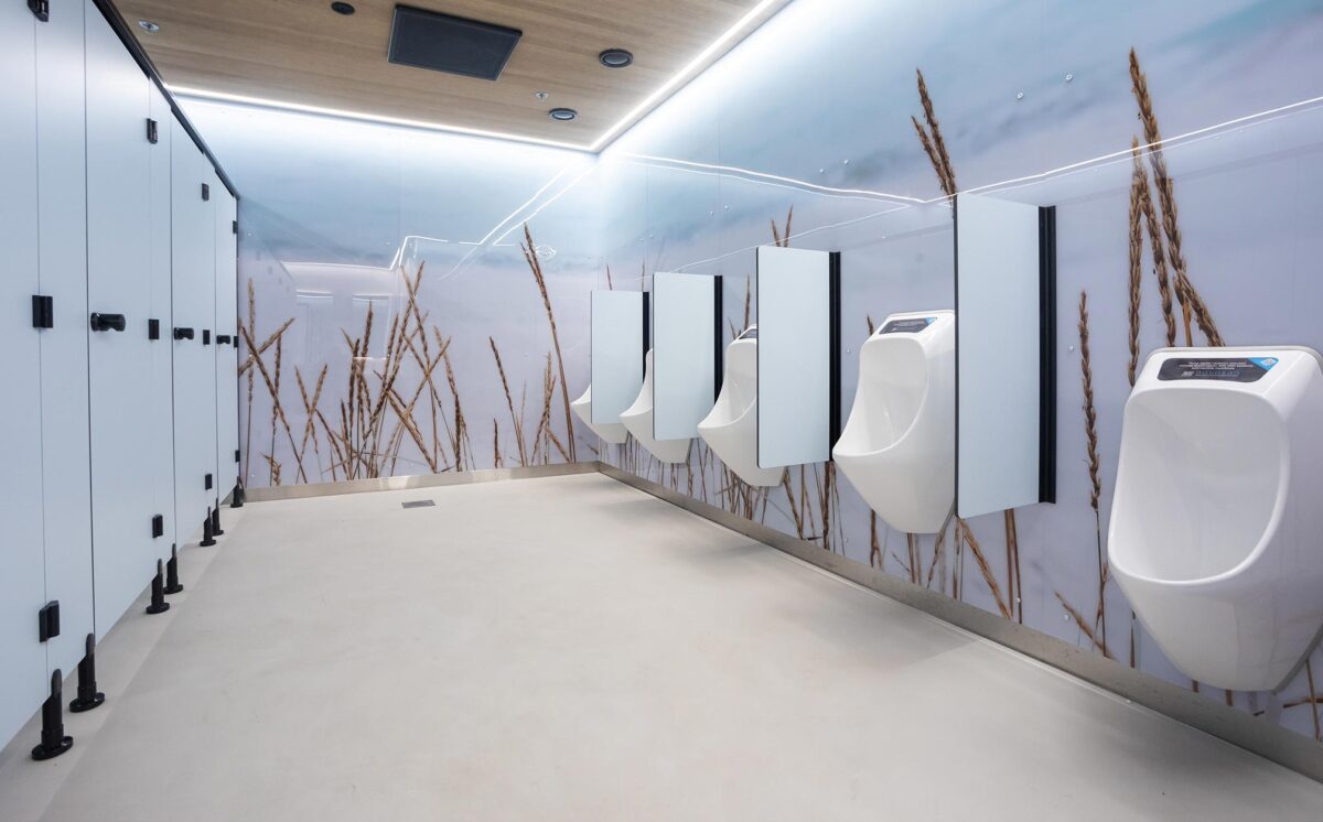 Sinisen sävyisessä miesten-WC:ssä on neljä vedetöntä pisuaaria väliseinineen ja eriöt. WC-tilan seinät ovat luontoaiheiset: viljatähkät nousevat kuvassa lattiasta kattoon ja taustalla näkyvät taivaan siniset ja violetit sävyt.