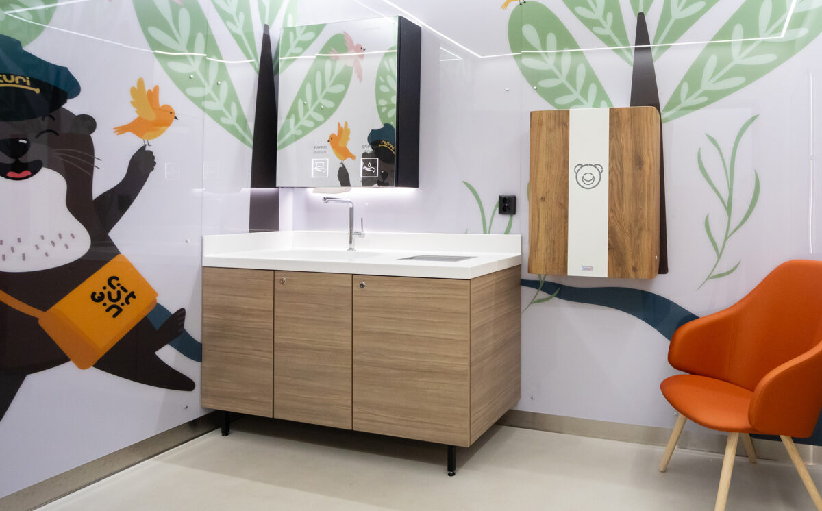 Lastenhoitohuoneessa on: seinään kiinnitetty vauvan hoitopöytä, peilikaappi, hana, allaskaappi ja oranssi nojatuoli. WC-tilan seinät ovat vaaleat ja niihin on kuvitettu hymyilevä saukko, lintu ja luontoa.