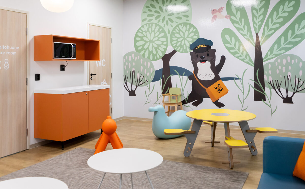Lasten leikkitila, jossa on värikäs sisustus ja kuvitettu seinämaalaus saukkohahmosta metsän keskellä. Huoneessa on oranssi kaappi mikroaaltouunilla, keltainen pöytä kiinteillä istuimilla sekä lapsille suunnattuja kalusteita, kuten vaaleansininen keinulelu ja oranssi leikkikoira. Ovet on merkitty teksteillä WC ja lastenhoitohuone suomeksi ja englanniksi.