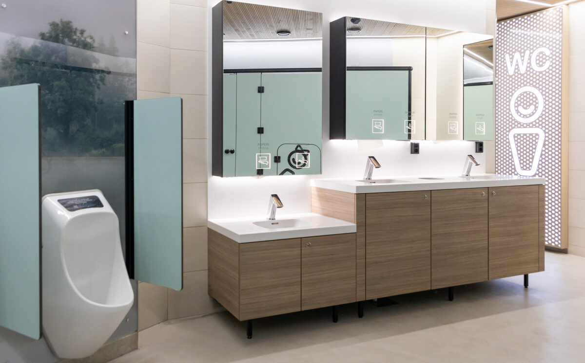 Moderni WC-tila, jossa automaattihanat, puupintaiset allaskaapit, valaistut peilit ja urinaali vihreillä väliseinillä. Oikealla sisäänkäynnissä on suurikokoinen valaistu miesten WC-symboli.