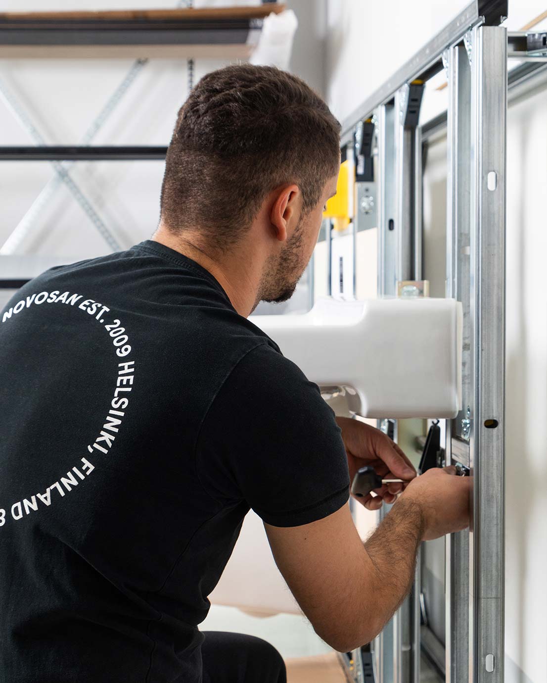 Mies asentaa pesuallasta WC-tilaelementtiin. Hänellä on musta paita, jossa lukee “NOVOSAN EST. 2009 HELSINKI, FINLAND”.