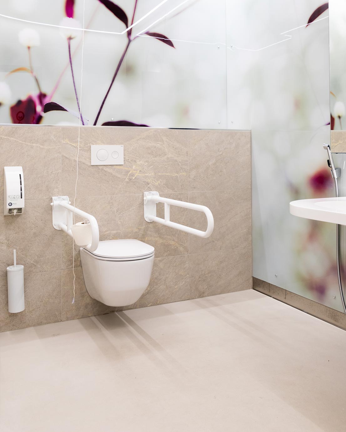 WC-tila, jossa on valkoinen seinä-WC-istuin ja sen molemmin puolin valkoiset kääntyvät tukikahvat. Taustaseinässä on kuvapinta, joissa on kuvituksena kukkia. WC-paperirulla on kiinnitetty vasemmanpuoleiseen tukikahvaan. Seinässä on myös annostelija ja valkoinen WC-harja telineessään. Lattia on vaalea.