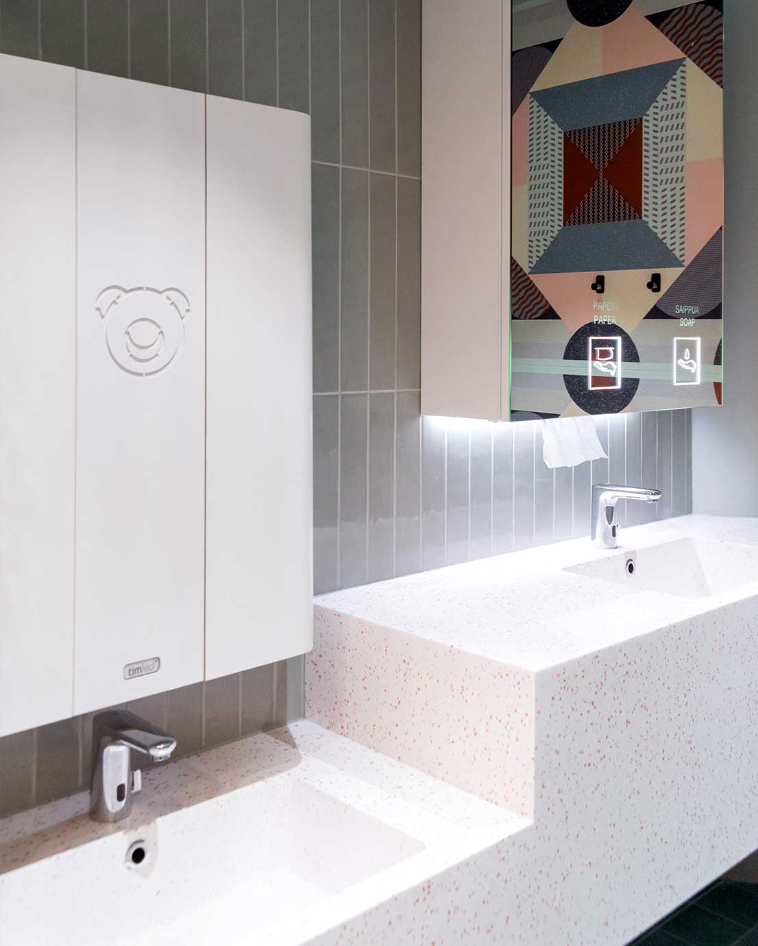 Novosan Timkid valkoinen lastenhoitopöytä julkisiin WC-tiloihin. Hoitopöytä on asennettu seinään hanan ja käsienpesualtaan yläpuolelle. Vauvan hoitopöydän vieressä on valaistu peilikaappi.