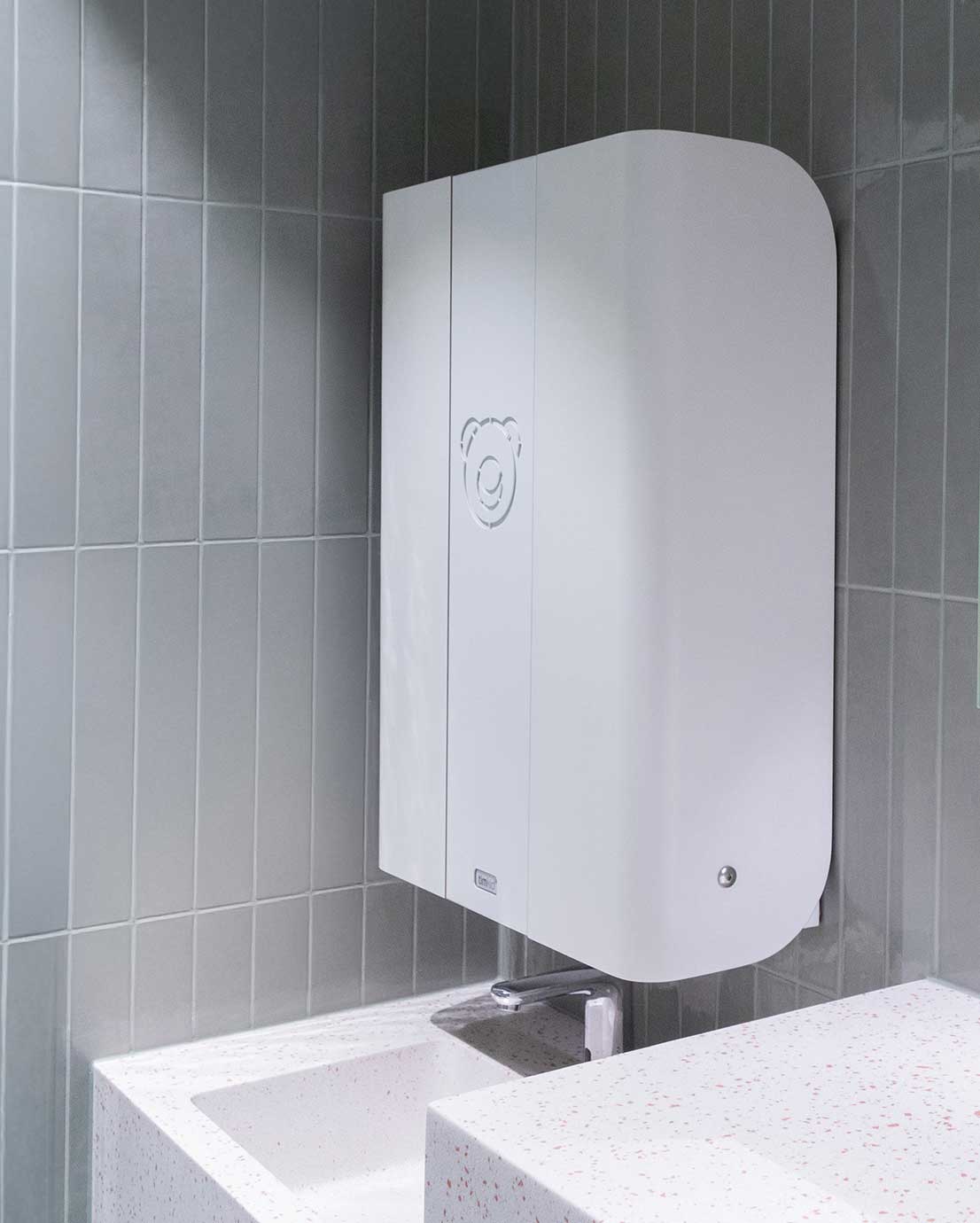 Novosan Timkid valkoinen lastenhoitopöytä julkisiin WC-tiloihin. Vauvan hoitopöydässä on pyöristetyt kulmat. Hoitopöytä on asennettu seinään hanan ja käsienpesualtaan yläpuolelle.