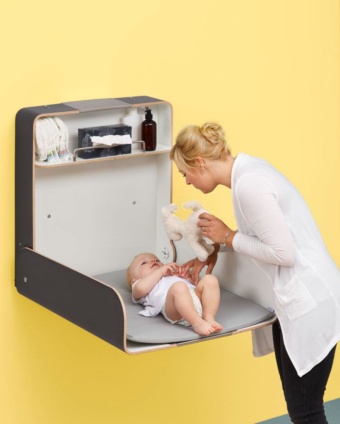 Nainen hoitaa vauvaa Novosan Timkid KAWAmaxi-hoitopöydällä. Hoitopöytä avattuna, taustalla lämmin keltainen seinä. Kuvassa korostuu tilava hoitopinta ja käytännön helppous.